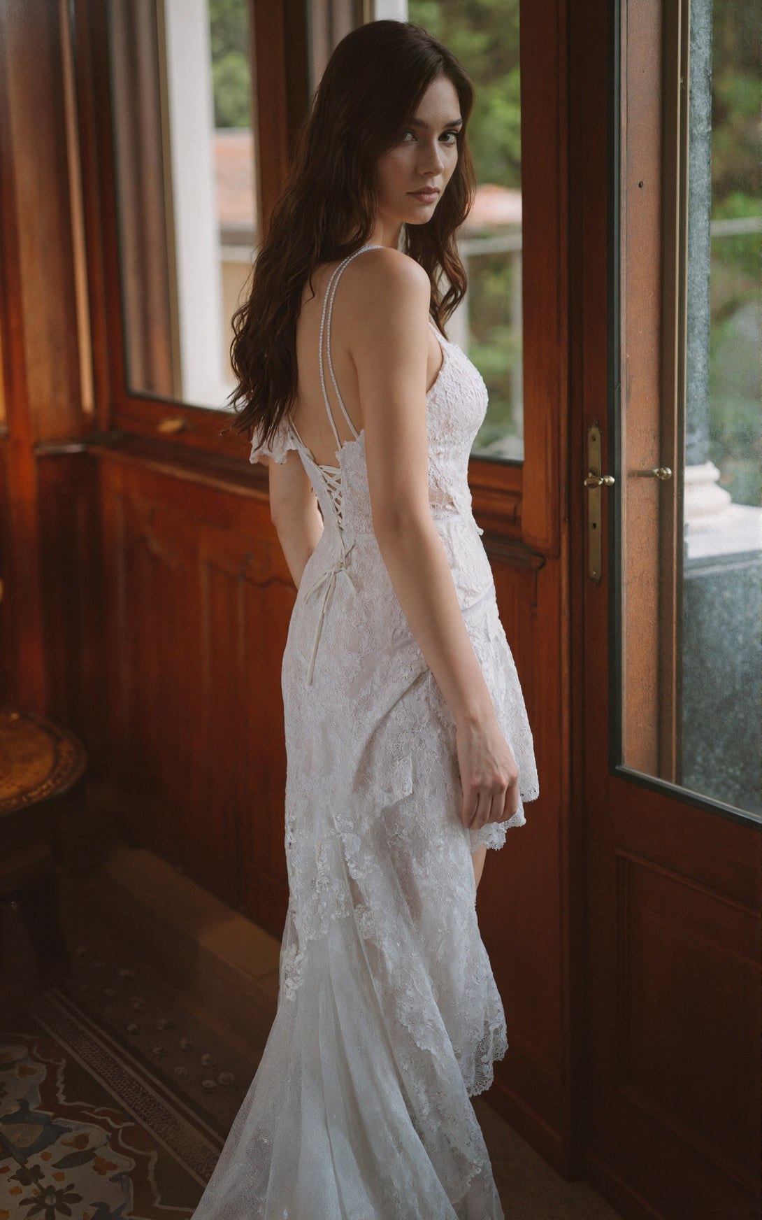 Woman in a white lace dress standing in a room with wooden walls and a door.