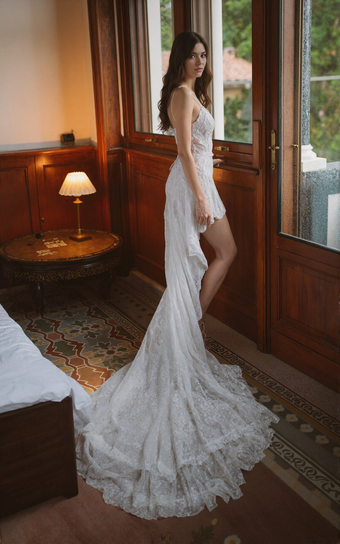 Woman in a white lace wedding dress standing in a room with wooden paneling and a lamp.